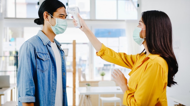 Asia female receptionist conducting wear protective face mask use infrared thermometer checker or temperature gun on customer's forehead before enter office. Lifestyle new normal after corona virus.