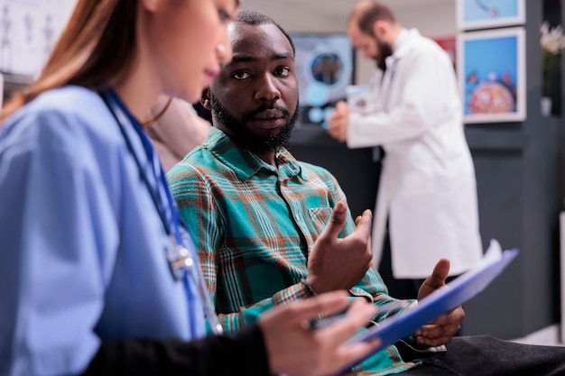 Asian assistant explaining diagnosis to male patient in hospital reception, doing checkup consultation in waiting area. Young man and specialist talking about healthcare treatment at clinic.