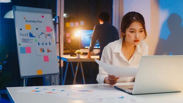 Asian businessman turn off computer and say goodbye his colleague who still working when he get off work after finish working overtime in small modern home office night. Coworker partnership concept.