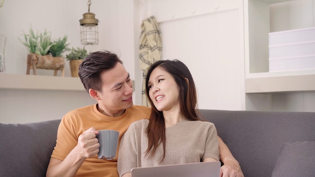 Asian couple using laptop and drinking warm cup of coffee in living room at home