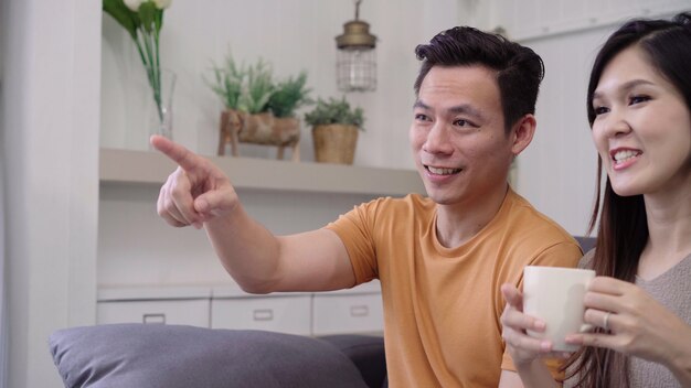 Asian couple watching TV and drinking warm cup of coffee in living room at home, sweet couple enjoy 