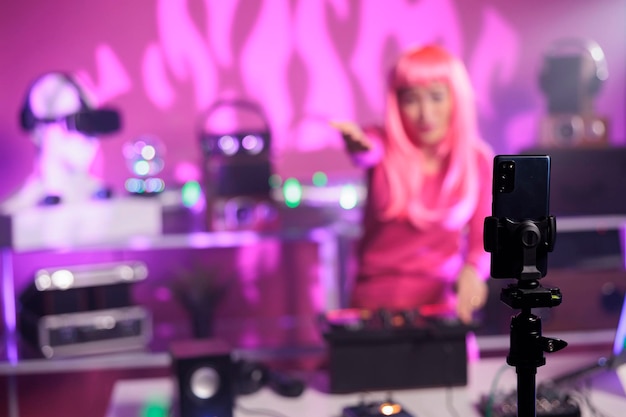 Free photo asian dj with pink hair performing techno music using professional mixer console while recording process with mobile phone camera. smiling artist playing song during night time in club