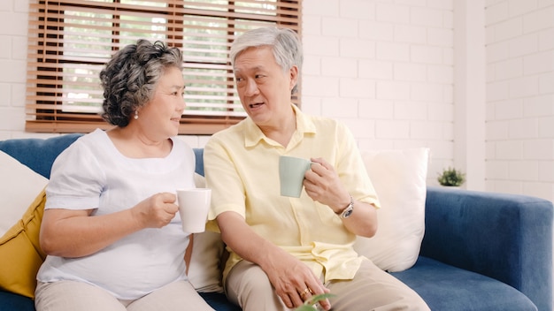 Asian elderly couple drinking warm coffee and talking together in living room at home, couple enjoy love moment while lying on sofa when relaxed at home. 