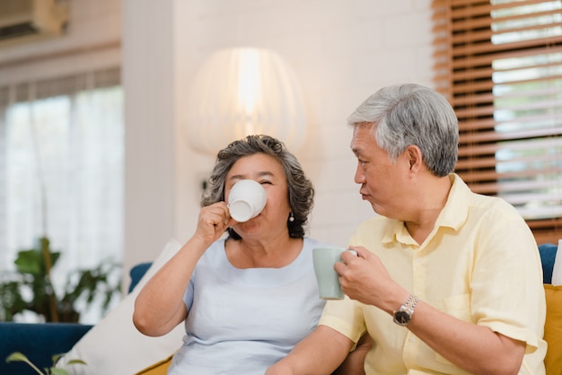 Free photo asian elderly couple drinking warm coffee and talking together in living room at home, couple enjoy love moment while lying on sofa when relaxed at home