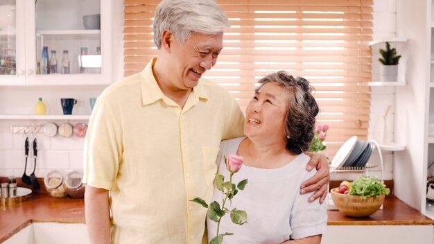 Asian elderly couple feeling happy smiling and holding flower and looking to camera while relax in kitchen at home. Lifestyle Senior family enjoy time at home concept. Portrait looking at camera.