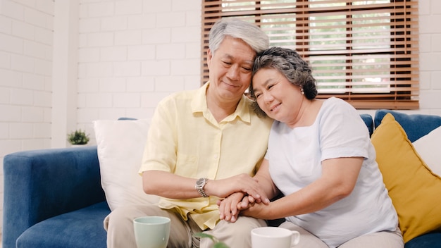 Asian elderly couple holding their hands while taking together in living room, couple feeling happy share and support each other lying on sofa at home. 