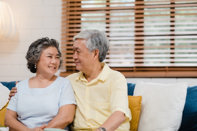 Asian elderly couple holding their hands while taking together in living room, couple feeling happy share and support each other lying on sofa at home. 