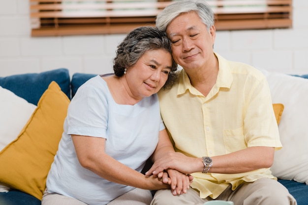 Free photo asian elderly couple holding their hands while taking together in living room, couple feeling happy share and support each other lying on sofa at home.