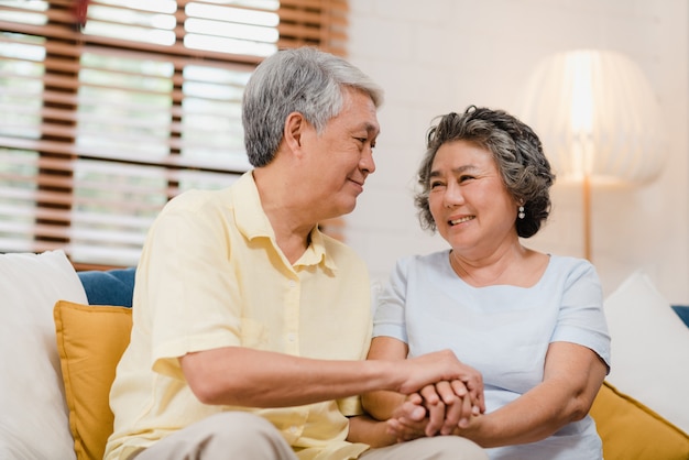 Asian elderly couple holding their hands while taking together in living room, couple feeling happy share and support each other lying on sofa at home. 