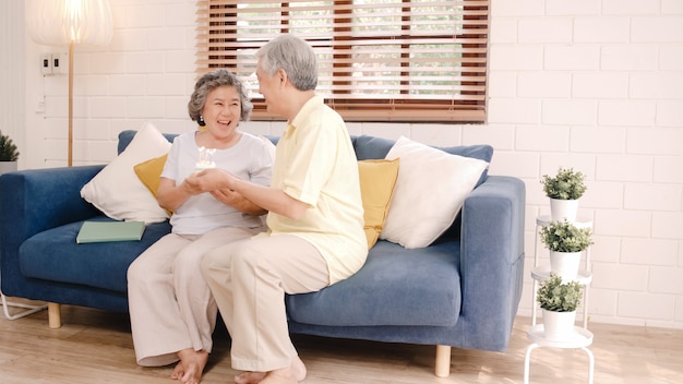 Asian elderly couple man holding cake celebrating wife's birthday in living room at home. Japanese couple enjoy love moment together at home.