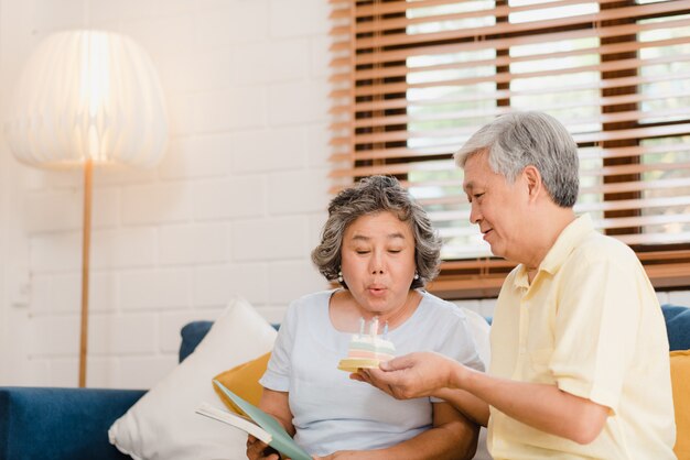 Asian elderly couple man holding cake celebrating wife's birthday in living room at home. Japanese couple enjoy love moment together at home. 