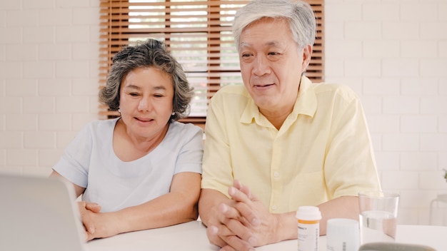 Free photo asian elderly couple using laptop conference with doctor about medicine information in living room, couple using time together while lying on sofa at home.