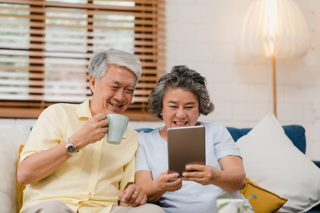 Asian elderly couple using tablet and drinking coffee in living room at home, couple enjoy love moment while lying on sofa when relaxed at home. 