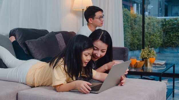 Asian family enjoy their free time relax together at home. Lifestyle mom and daughter using laptop watch movie on internet, dad watch TV in living room in modern home .