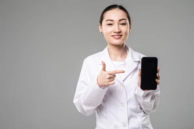 Asian female doctor smiling and showing a blank smart phone screen isolated on a white wall