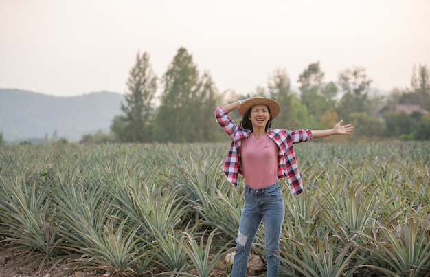 Asian female farmer see growth of pineapple in farm, Young pretty farmer woman standing on farmland with arms raised up joyful elated happiness.