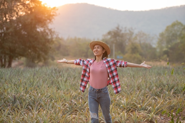 Asian female farmer see growth of pineapple in farm, Young pretty farmer woman standing on farmland with arms raised up joyful elated happiness.