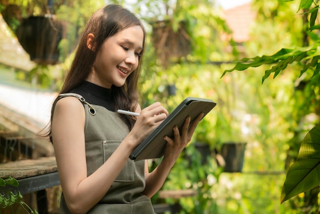 Asian female Woman gardener in apron working in a outdoor garden at home studioFemale Gardener using tablet computer for setting water drop system in greenhousemodern greenhouse farm ideas concept