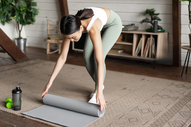 Free photo asian fitness girl finish trainingworkout at home rolling floor mat after exercises in living room