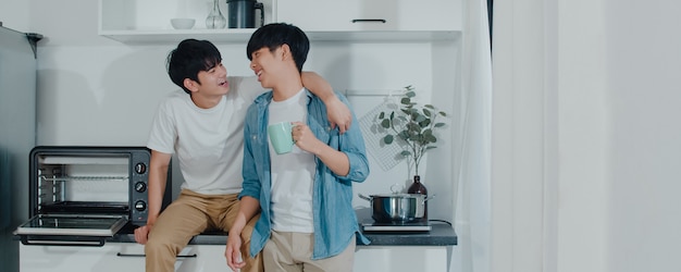 Asian gay couple drinking coffee, having a great time at home. Young handsome LGBTQ+ men talking happy relax rest together spend romantic time in modern kitchen at house in the morning .