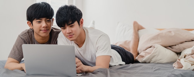 Asian Gay lgbtq men couple using computer laptop at modern home. Young Asia lover male happy relax rest together after wake up, watching movie lying on bed in bedroom at house in the morning .