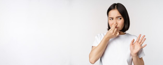 Asian girl looks disgusted rejecting product with bad smell shut nose from aversion and cringe standing against white background