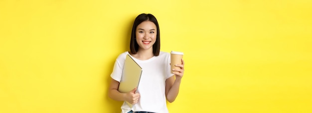 Free Photo asian girl student drinking coffee and holding laptop smiling at camera standing over yellow backgro