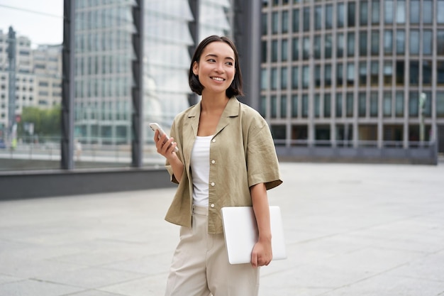 Free photo asian girl with laptop and smartphone standing on street of city centre smiling at camera