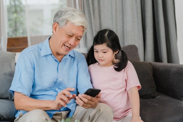 Free photo asian grandparents and granddaughter using mobile phone at home. senior chinese, grandpa and kid happy spend family time relax with young girl checking social media, lying on sofa in living room.