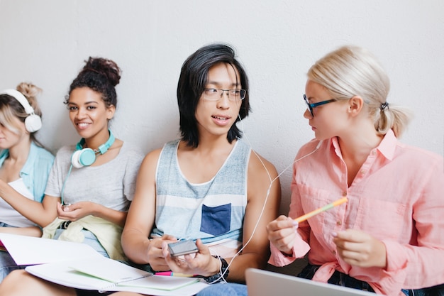 Free Photo asian guy with long hair discussing about new song with blonde female friend in glasses while black woman smiling. indoor portrait of students enjoying music and joking.