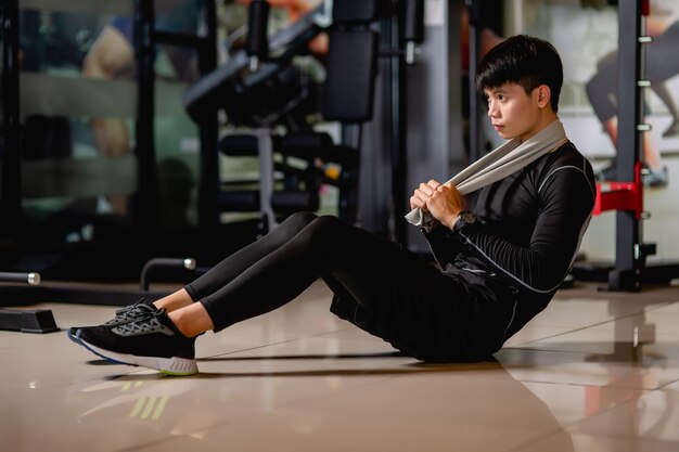 Asian handsome man wearing sportswear and smartwatch sitting on floor, sit-up to warm up muscle before workout at fitness gym, 