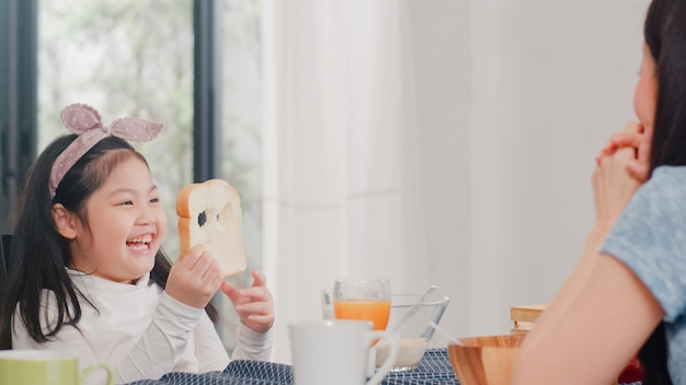 Asian Japanese family has breakfast at home. Asian daughter pick and play bread laughing smile with parents while eating corn flakes cereal and milk in bowl on table in modern kitchen in the morning.