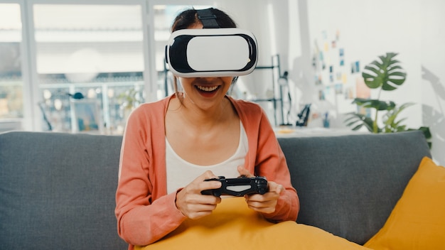 asian lady wear headset glasses of virtual reality play joystick game on couch in living room at house