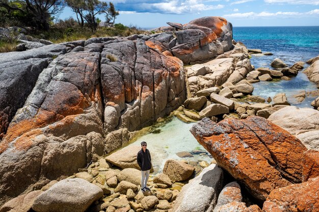 Asian male is posing for the camera while standing on big rocks next to a sea