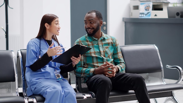 Asian medical assistant consulting male patient in medical clinic reception, sitting in waiting room lobby. Young man and nurse discussing about healthcare consultation and diagnosis.