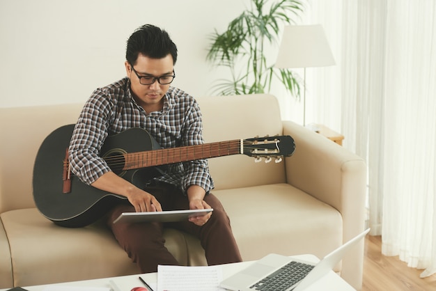 Asian musician sitting on couch at home with guitar and using tablet