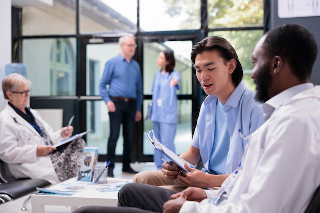 Asian patient holding clipboard filling medical report while discussing health care treatment with specialist medic during consultation in hospital waiting area. Diverse people standing in reception