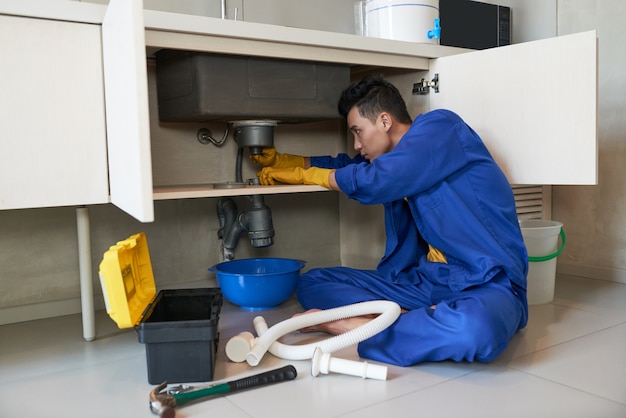 Asian plumber in blue overalls clearing blockage in drain