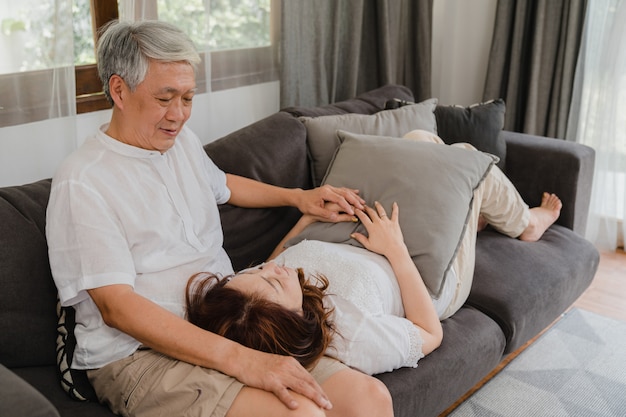 Asian senior couple relax at home. Asian Senior Chinese grandparents, husband happy smile hug lie down her wife lap while lying on sofa in living room at home concept.