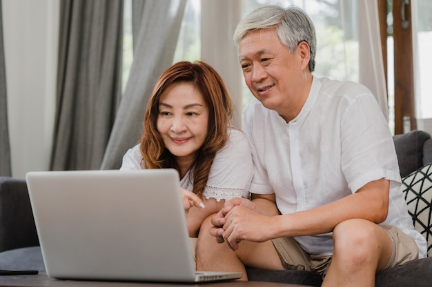 Free photo asian senior couple using laptop at home. asian senior chinese grandparents, surf the internet to check social media while lying on sofa in living room at home concept.