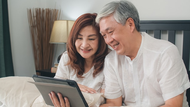 Free photo asian senior couple using tablet at home. asian senior chinese grandparents, video call talking with family grandchild kids while lying on bed in bedroom at home in the morning concept.