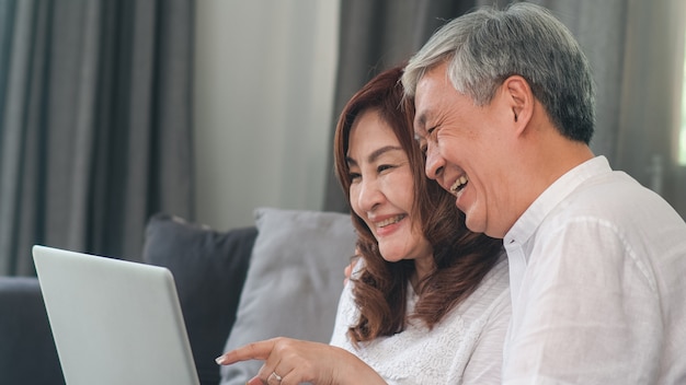 Free photo asian senior couple video call at home. asian senior chinese grandparents, using laptop video call talking with family grandchild kids while lying on sofa in living room at home concept.