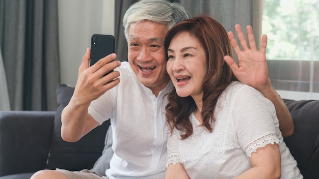 Asian senior couple video call at home. Asian Senior Chinese grandparents, using mobile phone video call talking with family grandchild kids while lying on sofa in living room at home concept.