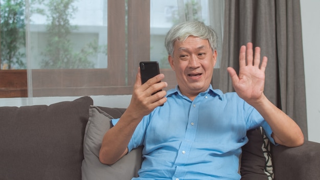 Free photo asian senior man video call at home. asian senior older chinese male using mobile phone video call talking with family grandchild kids while lying on sofa in living room at home concept.