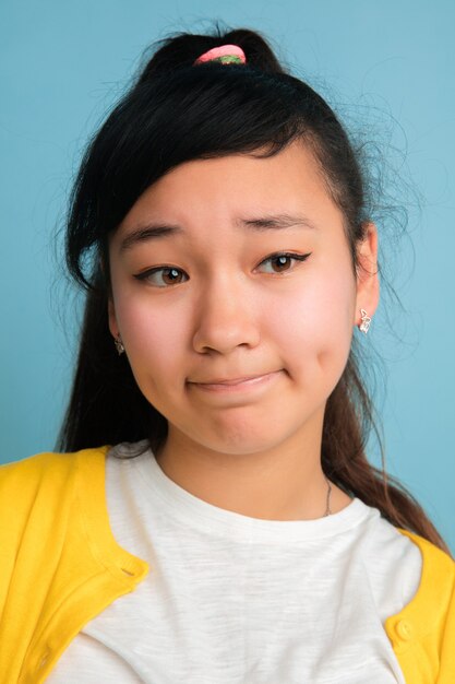 Asian teenager's close up portrait isolated on blue studio background. Beautiful female brunette model with long hair. Concept of human emotions, facial expression, sales, ad. Looks sad.