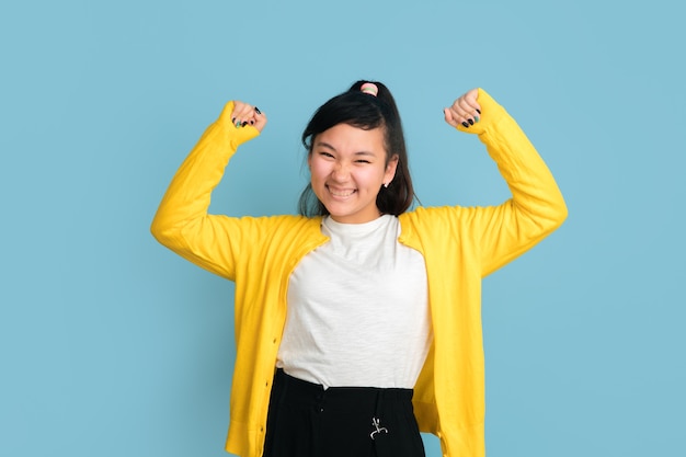 Asian teenager's portrait isolated on blue studio background. Beautiful female brunette model with long hair. Concept of human emotions, facial expression, sales, ad. Happy winning, bet concept.