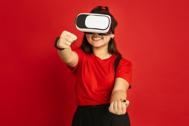 Asian teenager's portrait isolated on red studio background. Beautiful female brunette model with long hair in casual style. Concept of human emotions, facial expression, sales, ad. Plays VR-headset.