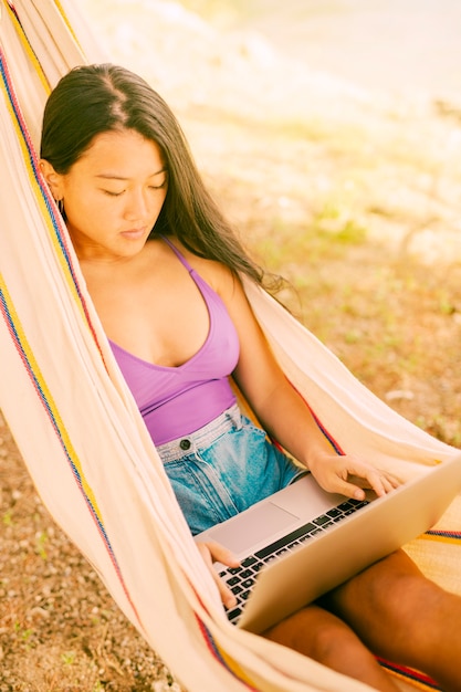 Free photo asian woman lying in hammock and working on laptop