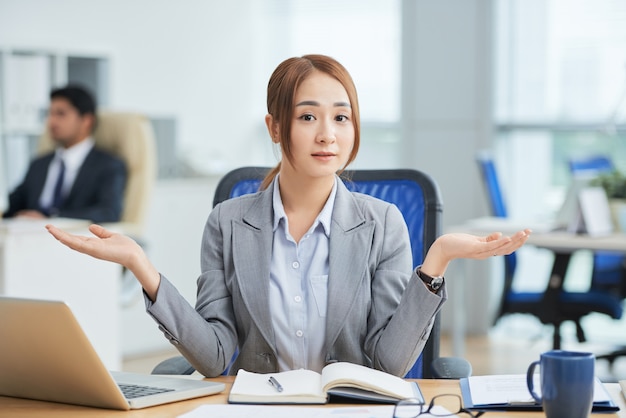 Asian woman sitting at desk in office and looking at camera with helpless hand gesture