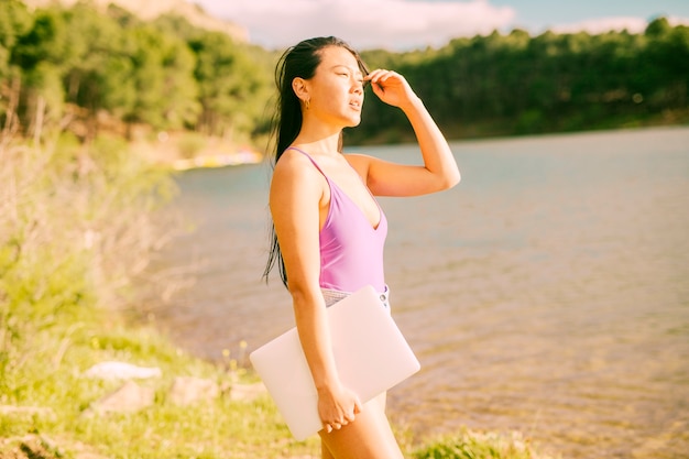 Free photo asian woman standing near lake and holding laptop